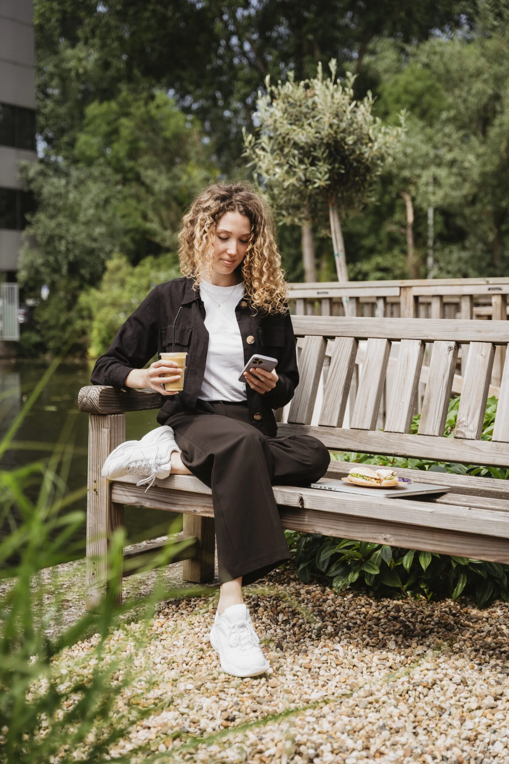 Vrouw zit op een houten bank in de tuin van HNK Amsterdam Schinkel, met een koffie en smartphone in de hand.