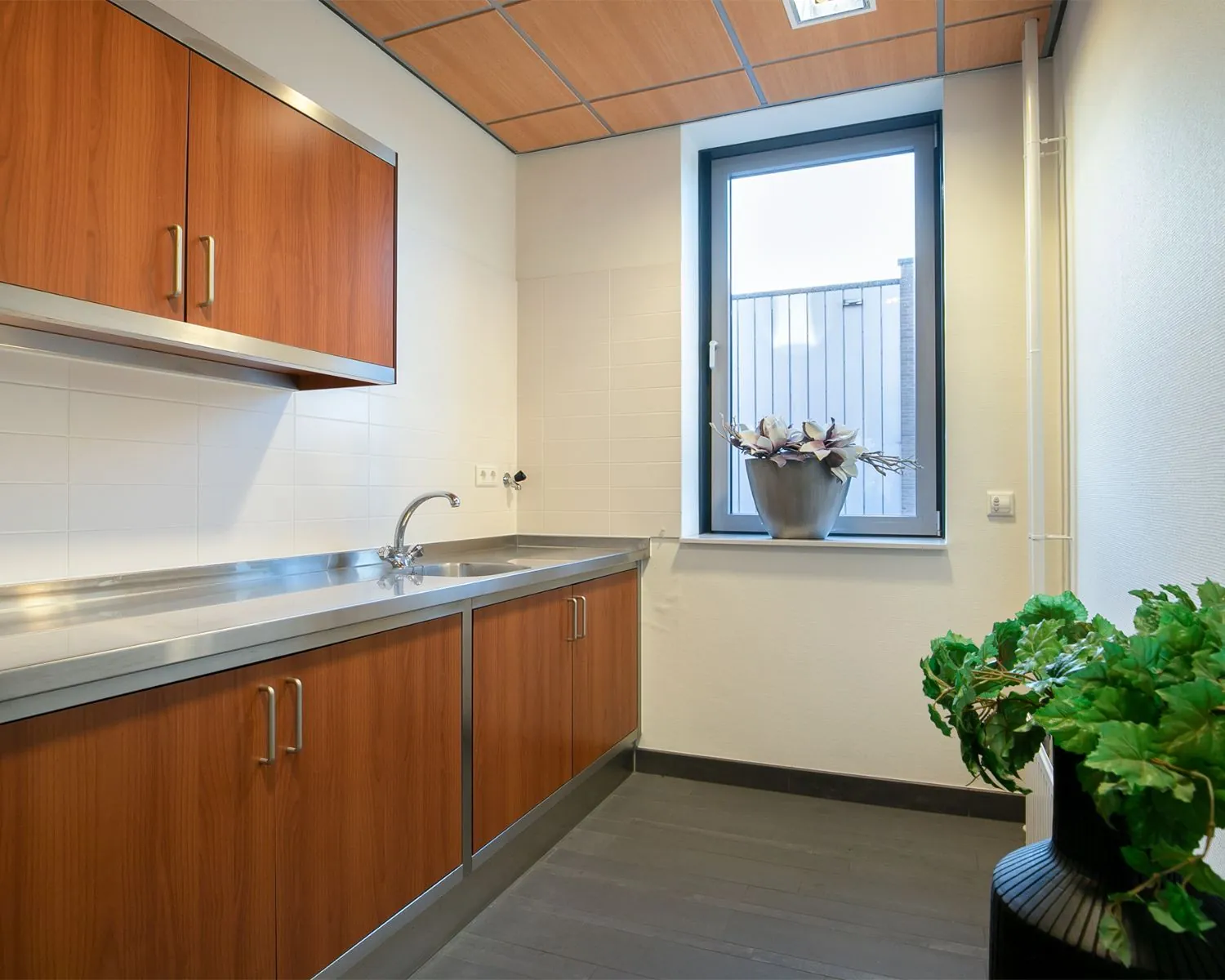 Kleine pantry met houten keukenkasten, rvs-aanrechtblad en een raam met planten, in kantoorruimte aan de Carlsonstraat 13 in Kampen.
