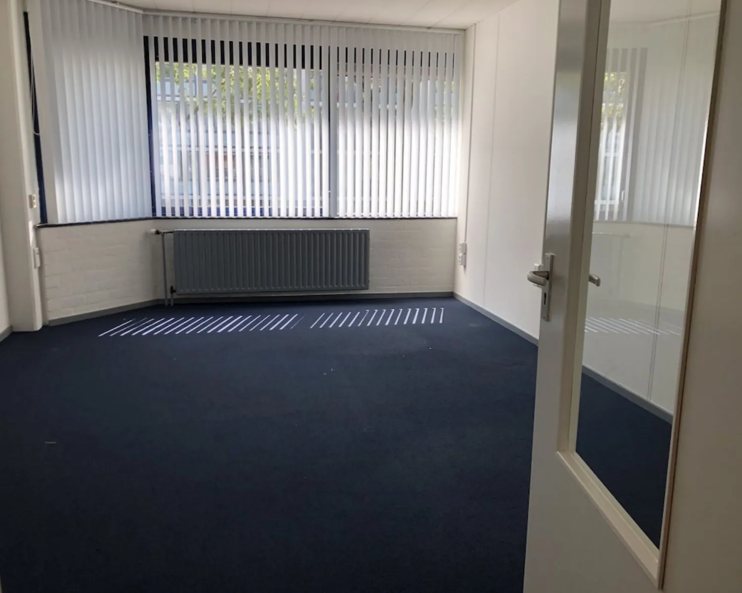 Leeg kantoor met donkerblauwe vloerbedekking en grote ramen met verticale lamellen aan de Hooilaan 1 in Breda.