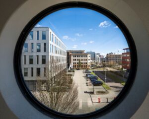 Zicht op moderne kantoorgebouwen en een parkeerplaats aan de Wegalaan 35 in Hoofddorp, gefotografeerd door een rond raam.