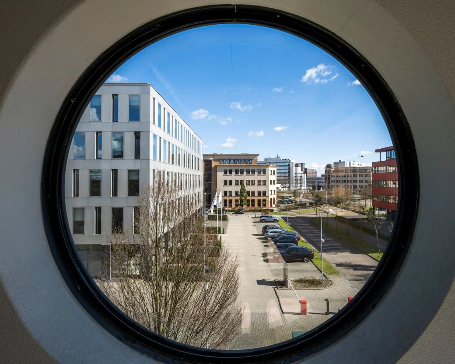 Zicht op moderne kantoorgebouwen en een parkeerplaats aan de Wegalaan 35 in Hoofddorp, gefotografeerd door een rond raam.