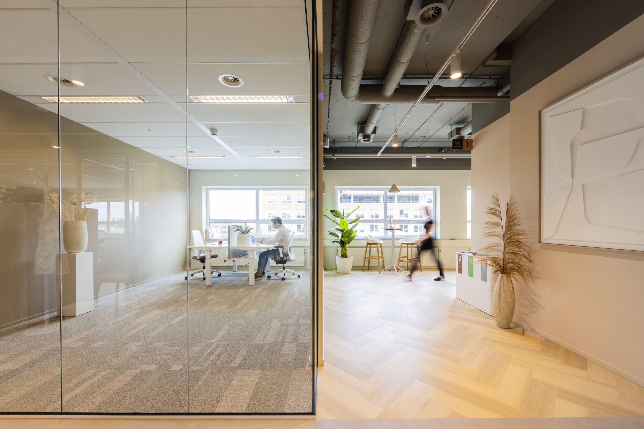 Een modern kantoor met een glazen wand, waar een persoon zit te werken aan een bureau en een ander persoon langsloopt naast hoge krukken en een grote plant.