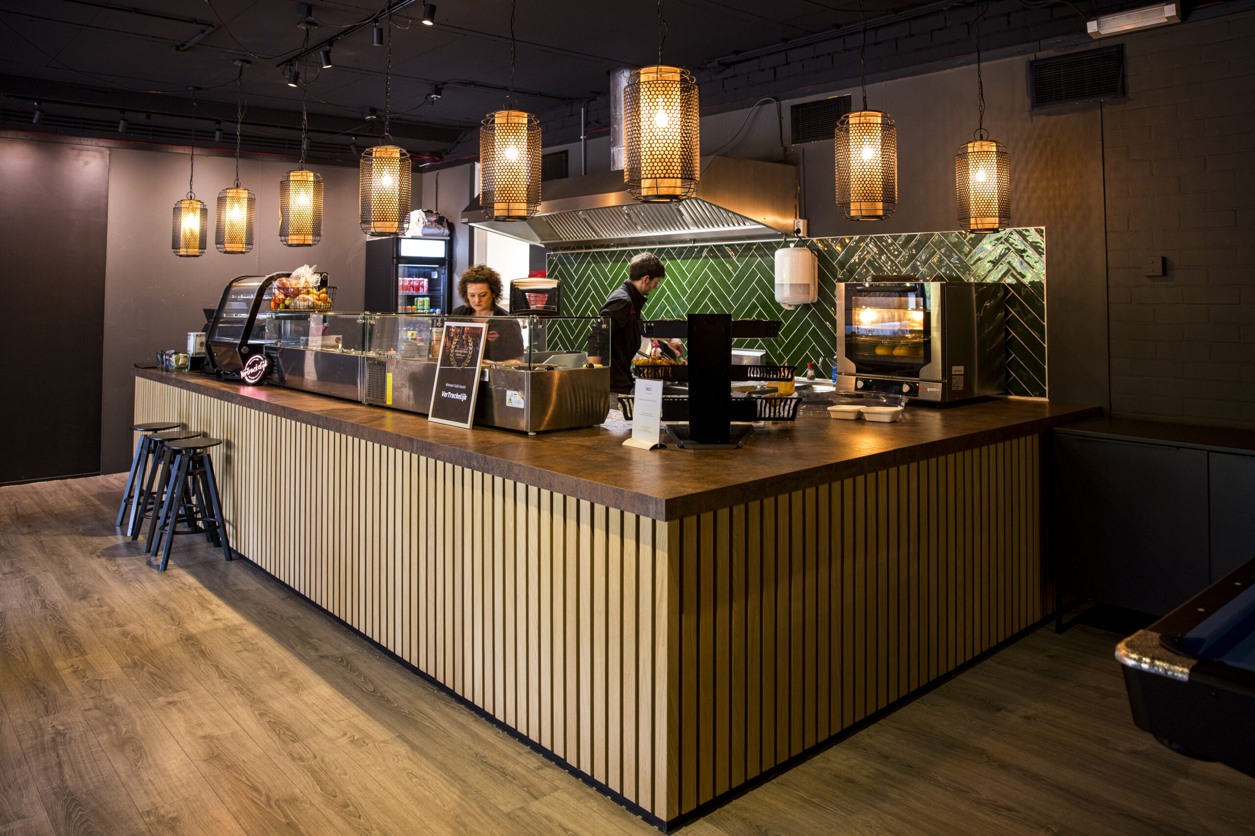 Een moderne restaurantbalie met sfeervolle hanglampen en personeel in de keuken bij Restaurant ELE_ALM.