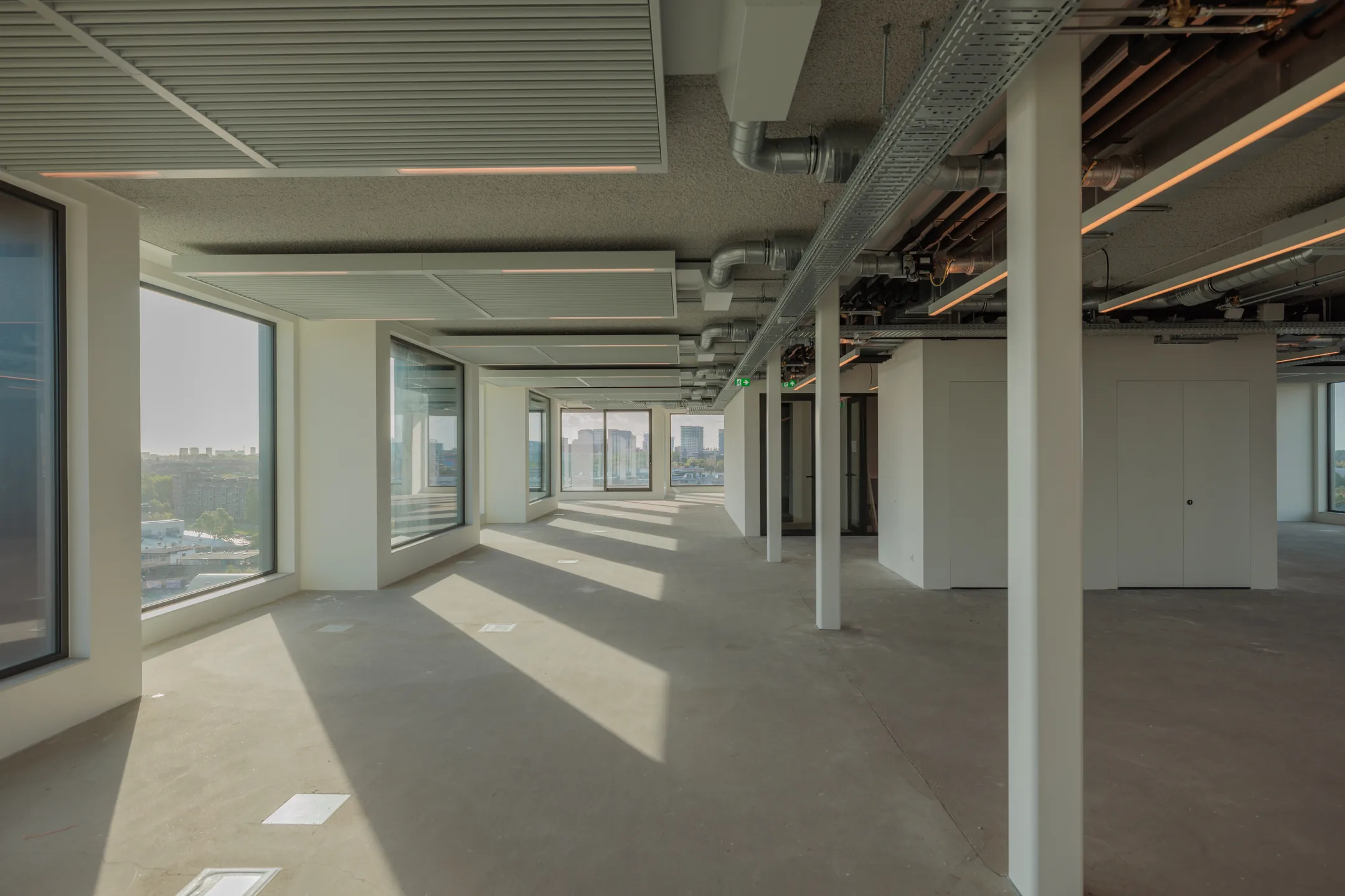 Een open, leeg kantoorruimte met grote ramen, plafonds voorzien van zichtbare ventilatiebuizen.