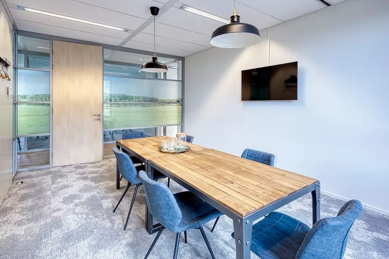 Moderne vergaderruimte aan de Mercatorlaan met houten tafel, zes blauwe stoelen, muurtelevisie en glazen wanden met natuurfoto.