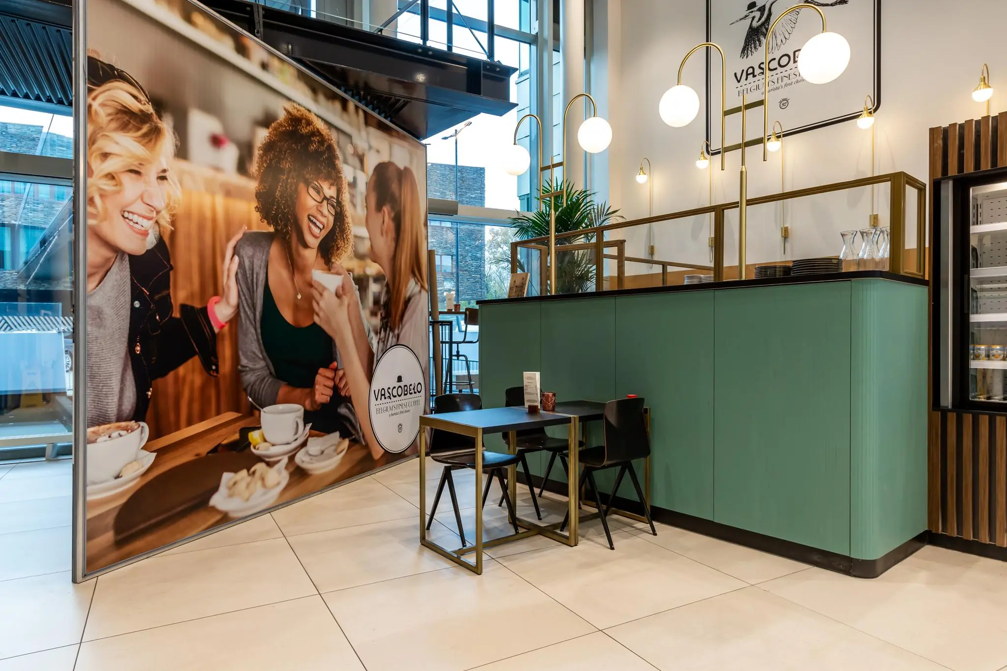 Moderne koffiezaak Vascobelo op het Herculesplein met stijlvolle zitplek en grote wandposter van lachende mensen aan tafel.