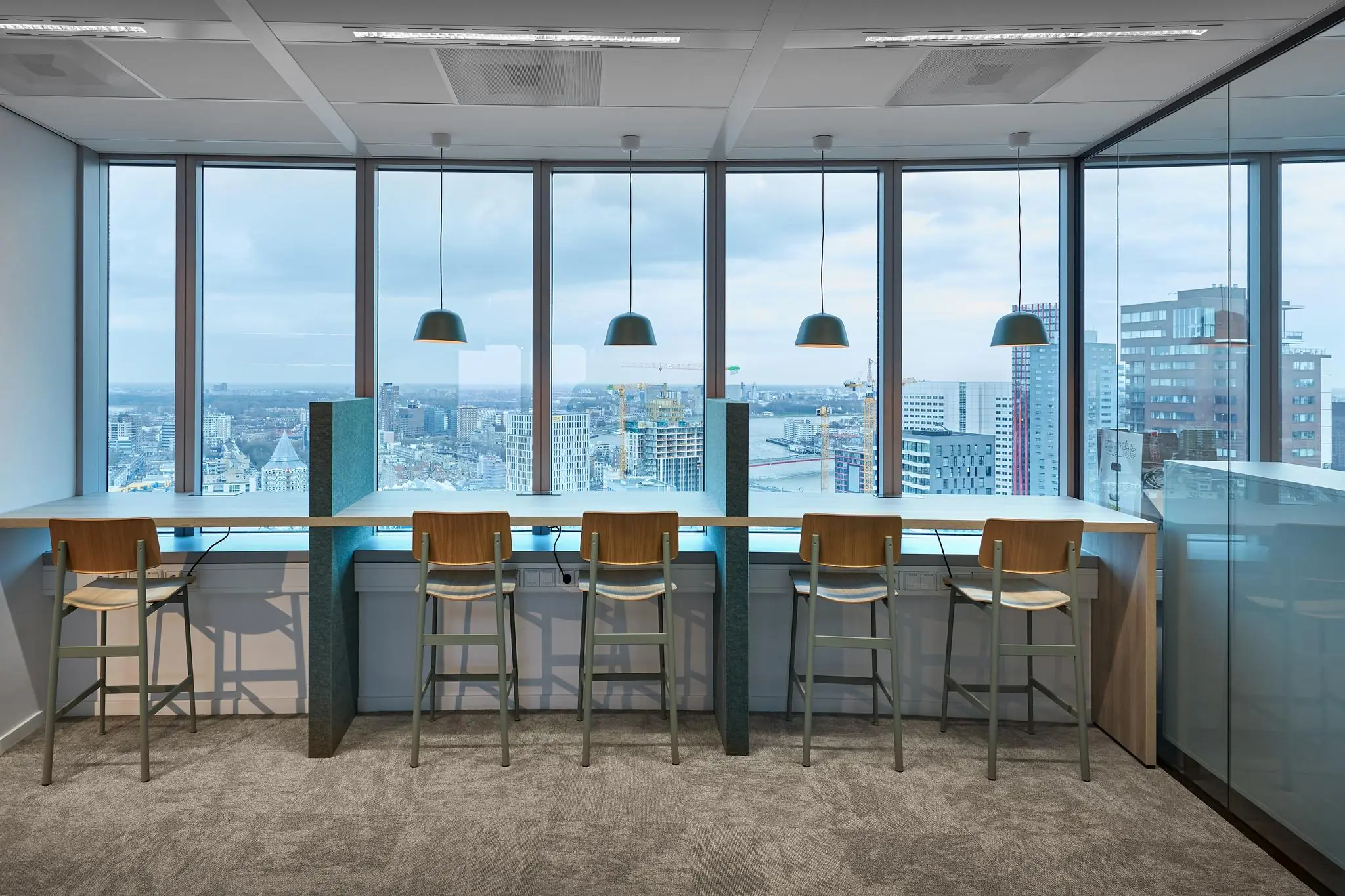 Moderne werkplek met hoge stoelen aan een lange tafel bij het raam, met uitzicht op de Rotterdamse wijk Blaak.