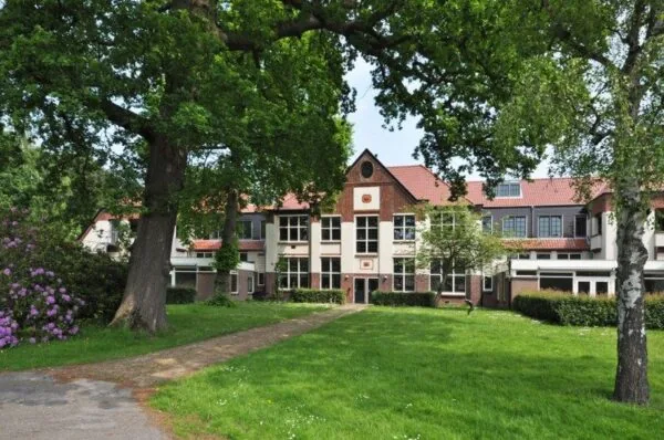 Voorgevel van een groot, traditioneel gebouw aan de Spechtlaan, omgeven door gras, bomen en bloeiende struiken.