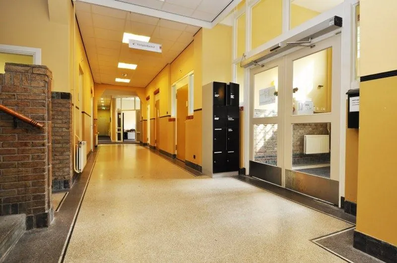 Binnenhal van een school of zorginstelling aan de Spechtlaan, met gele muren, lockers en zicht op meerdere deuren en gangen.