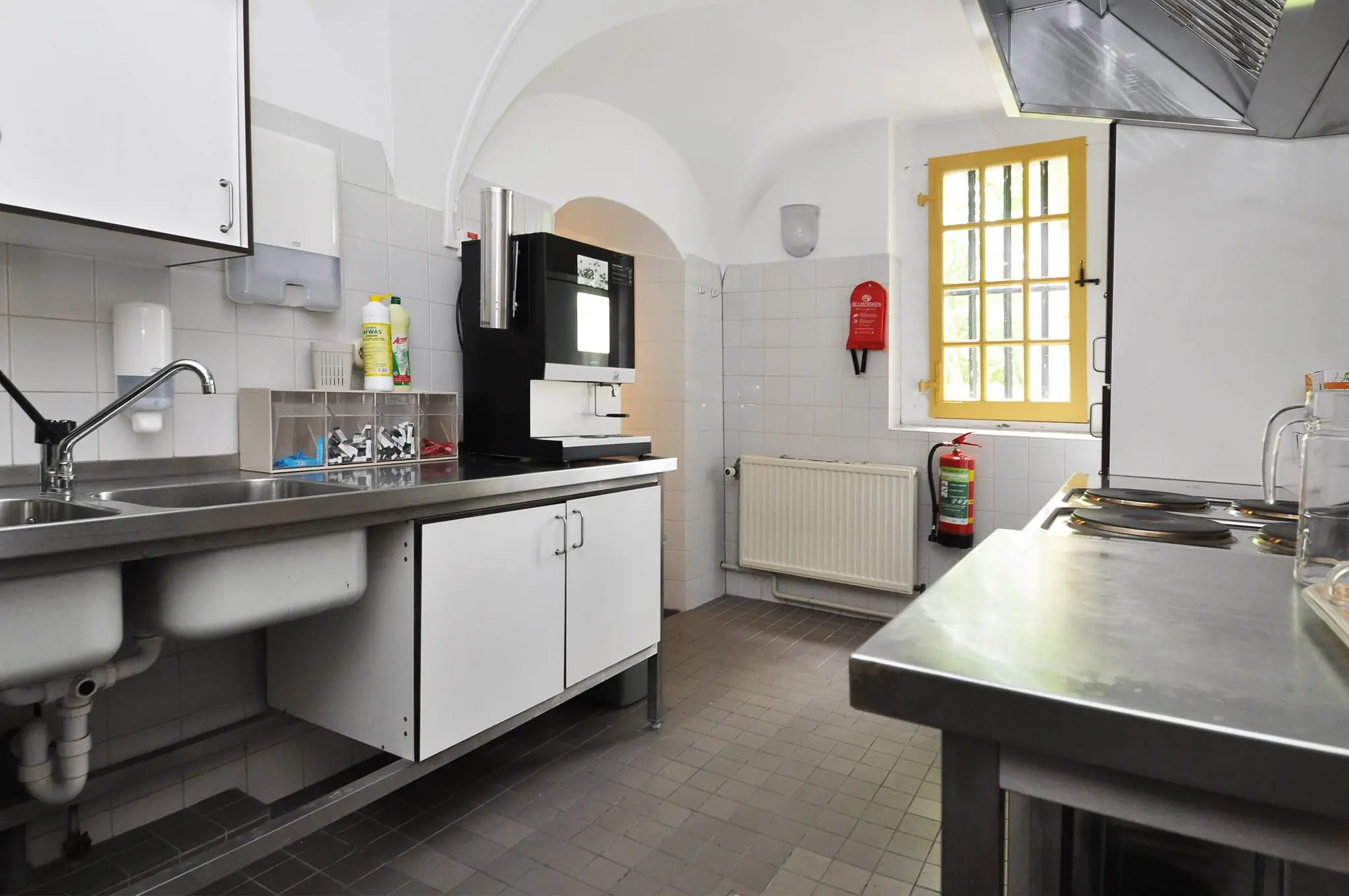Professionele keuken met roestvrijstalen werkbladen, dubbele spoelbak, koffieautomaat en gele raamkozijnen aan de Kasteellaan.