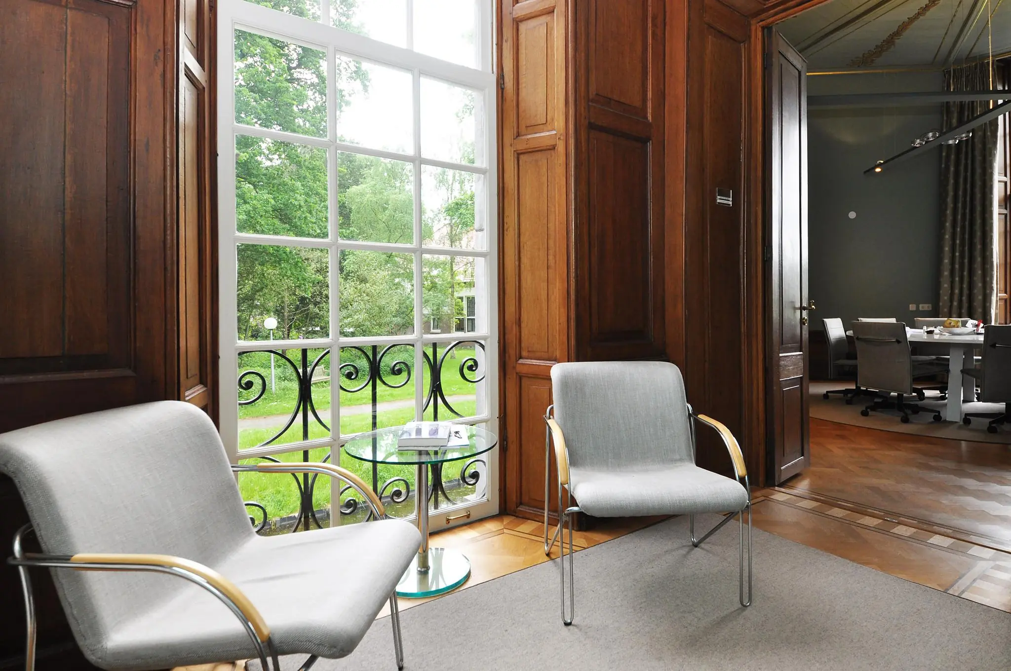 Wachtruimte met twee moderne stoelen, een glazen bijzettafel en uitzicht op een groene tuin via een groot raam in Kasteellaan.