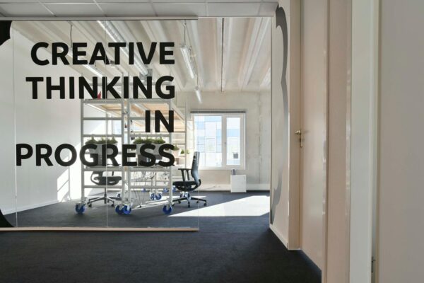 Glazen kantoormuur met de tekst "Creative thinking in progress" en zicht op een moderne werkruimte met bureaustoelen en plantenrekken.