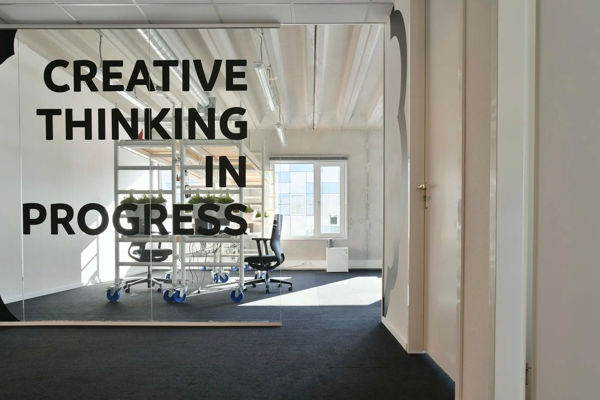 Glazen kantoormuur met de tekst "Creative thinking in progress" en zicht op een moderne werkruimte met bureaustoelen en plantenrekken.