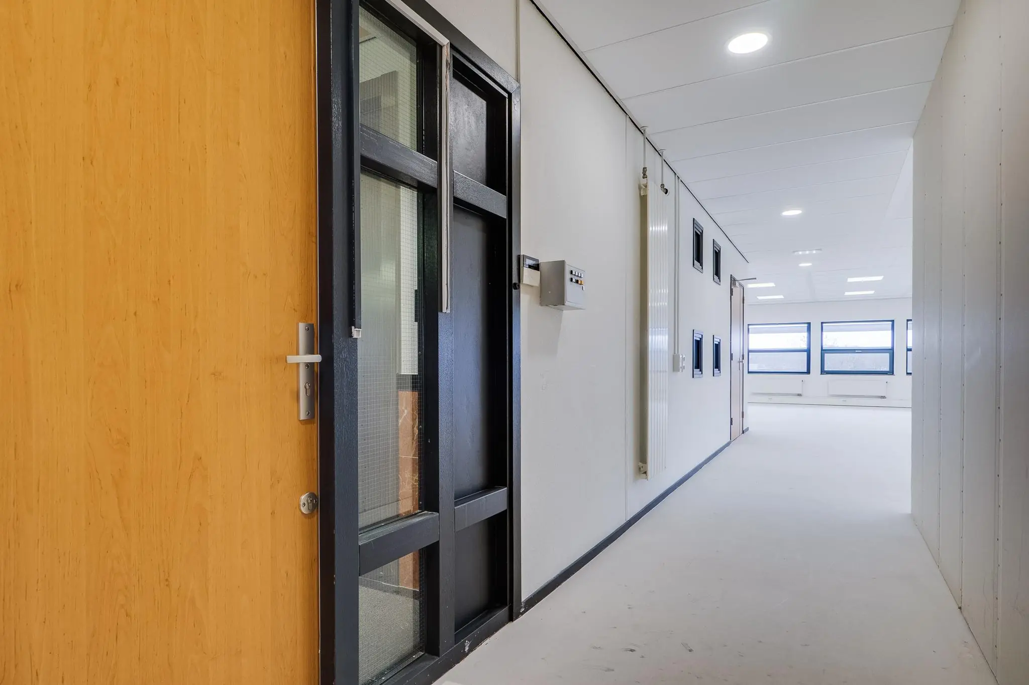 Licht, modern kantoorinterieur met een gang, houten deur en grote ramen aan het einde.