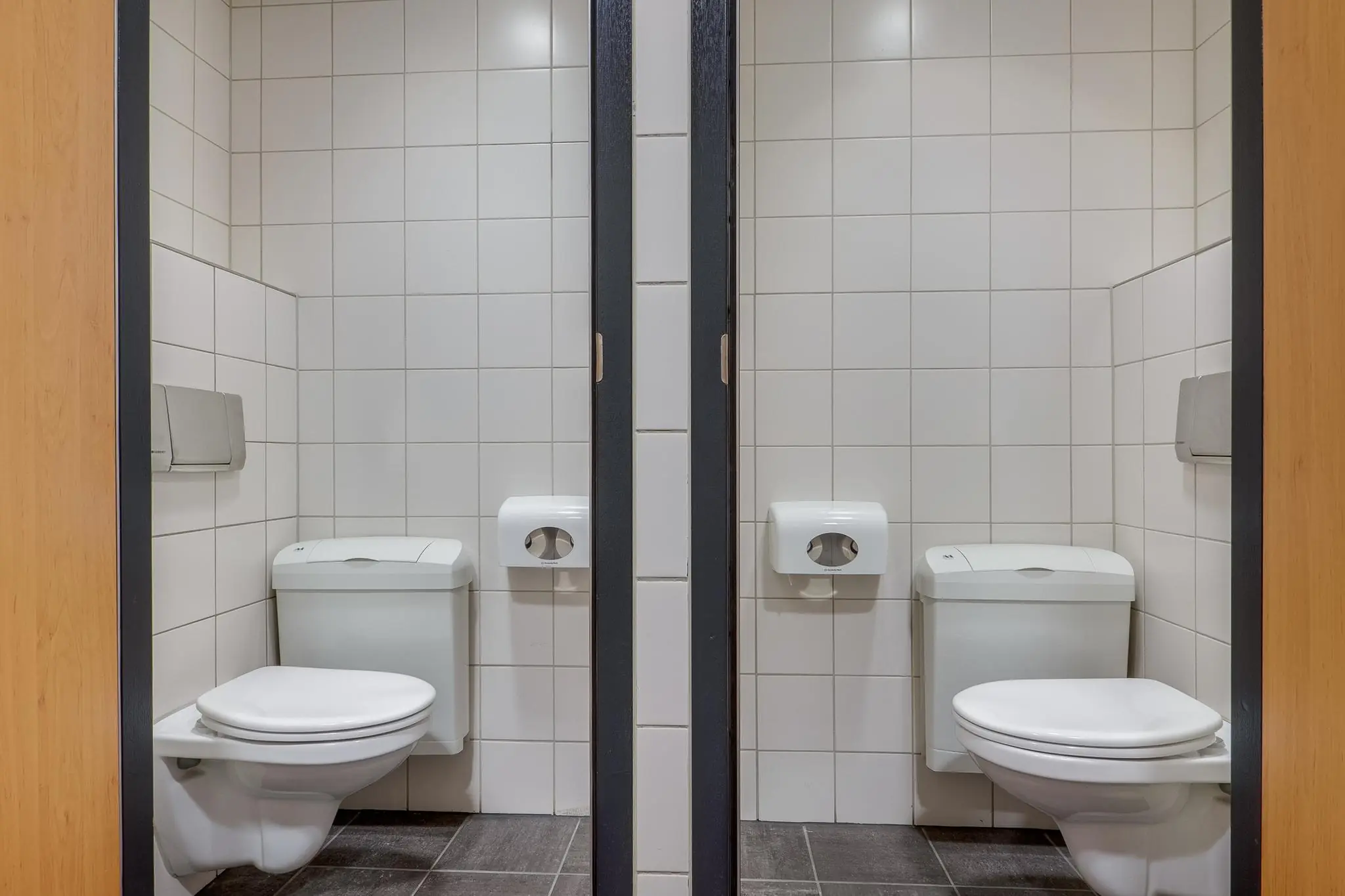 Twee naast elkaar gelegen toiletcabines met witte wandtegels en wandclosetten in een openbaar gebouw aan de Marconibaan.