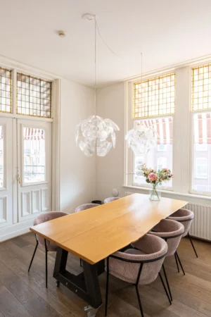 Een lichte eetkamer met een houten tafel, zes gestoffeerde stoelen, moderne hanglampen en een vaas bloemen bij de ramen met glas-in-lood details.