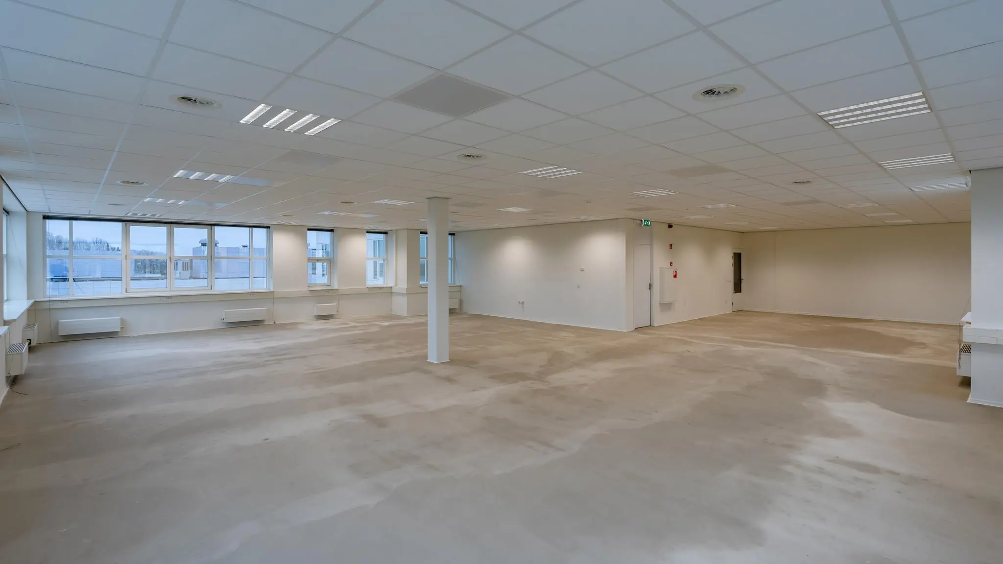 Lege kantooretage in het Rhijnspoorgebouw met systeemplafond, grote ramen en betonnen vloer.
