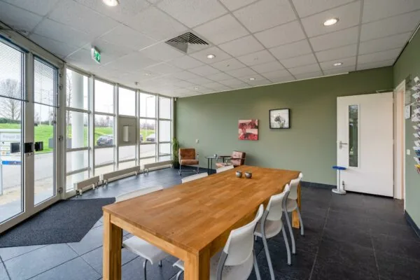 Moderne wachtruimte bij Rhijnspoor 251 met grote ramen, houten tafel, witte stoelen en twee leren fauteuils.