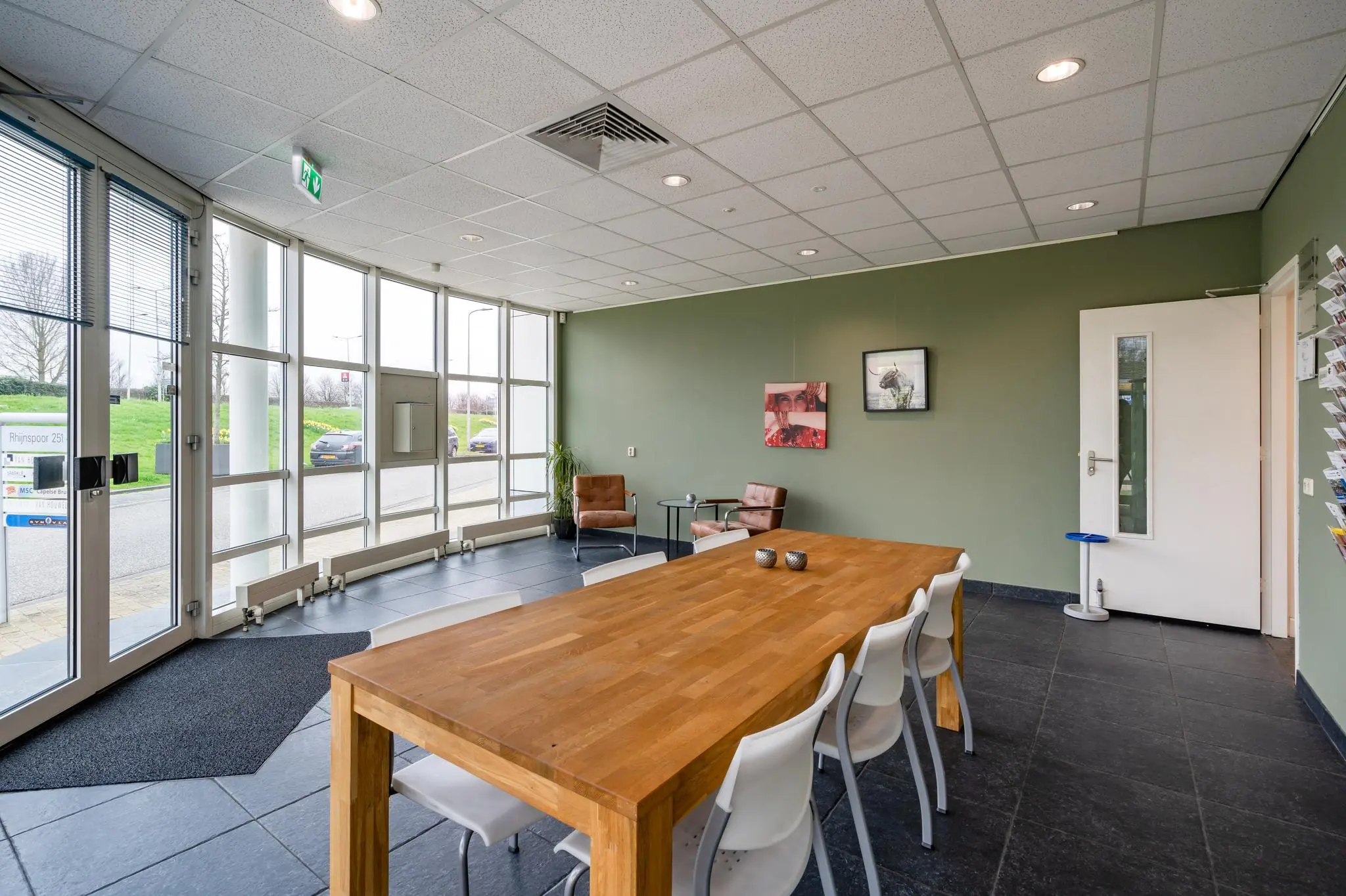 Moderne wachtruimte bij Rhijnspoor 251 met grote ramen, houten tafel, witte stoelen en twee leren fauteuils.