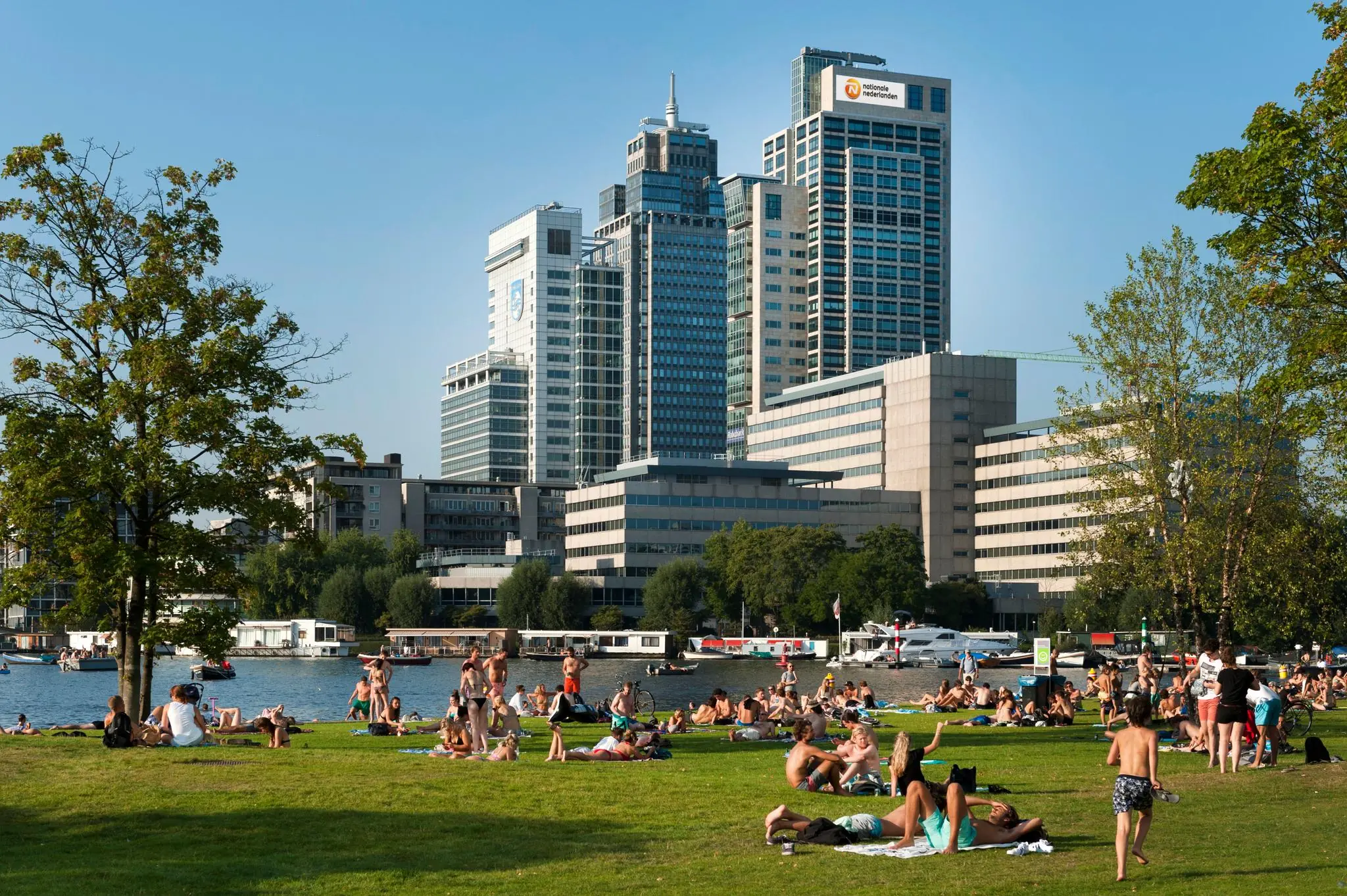 Mensen genieten van de zon in het gras bij het Amstelplein in Amsterdam, met kantoorgebouwen op de achtergrond.
