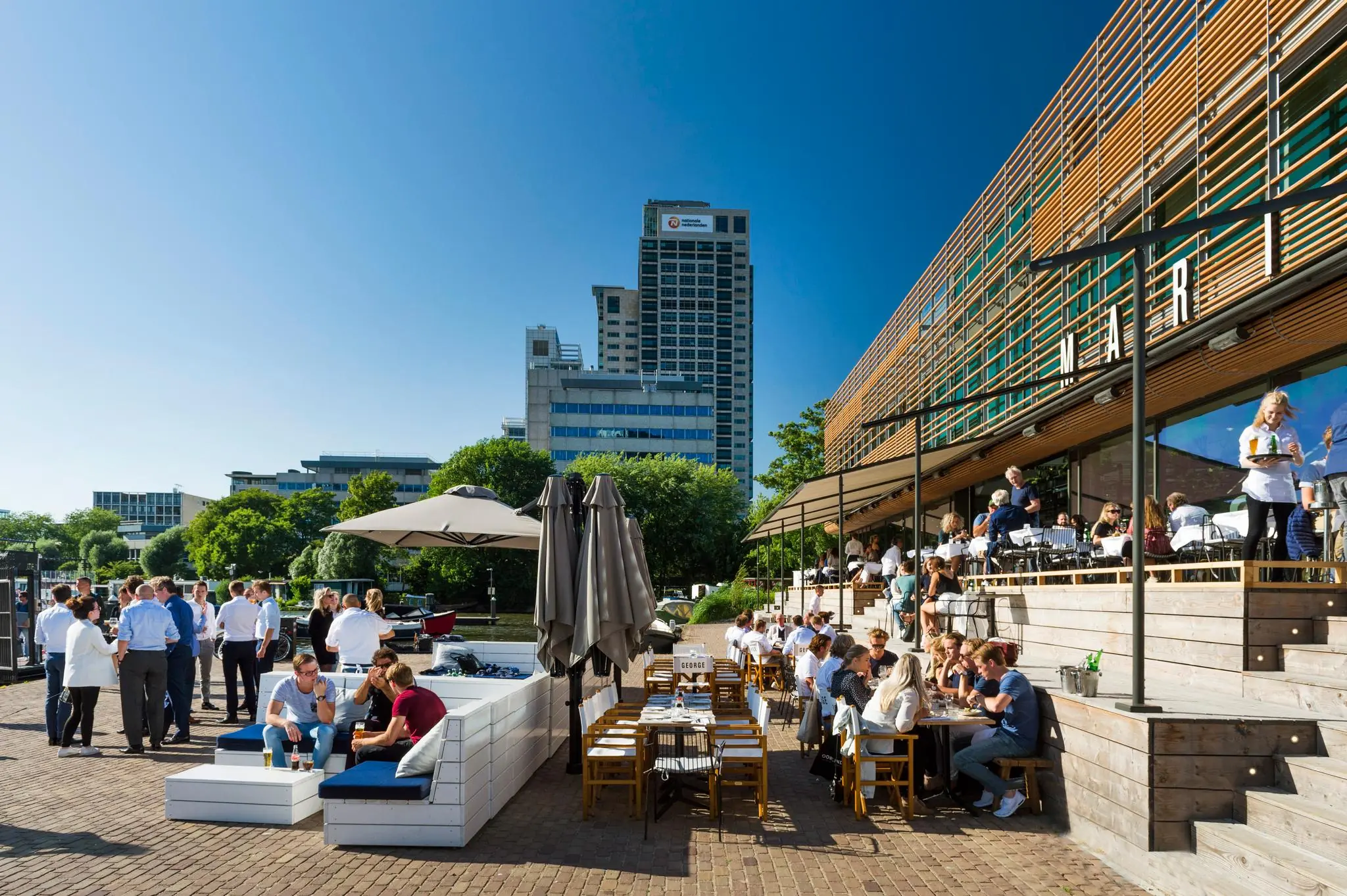 Terras vol mensen bij restaurant George Marina aan het Amstelplein in Amsterdam op een zonnige dag.