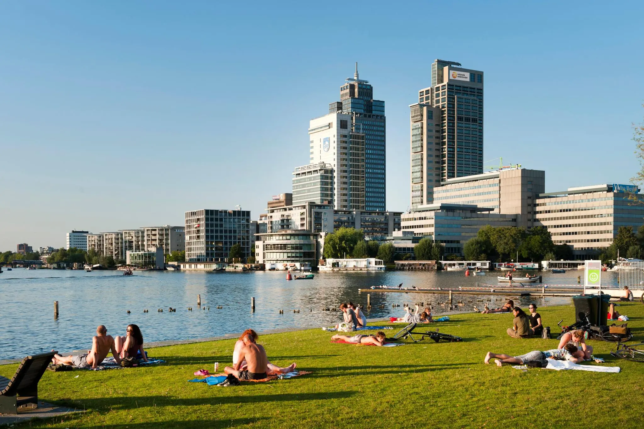 Mensen ontspannen op het gras bij de Amstel met de hoogbouw van Amstelplein op de achtergrond op een zonnige dag.