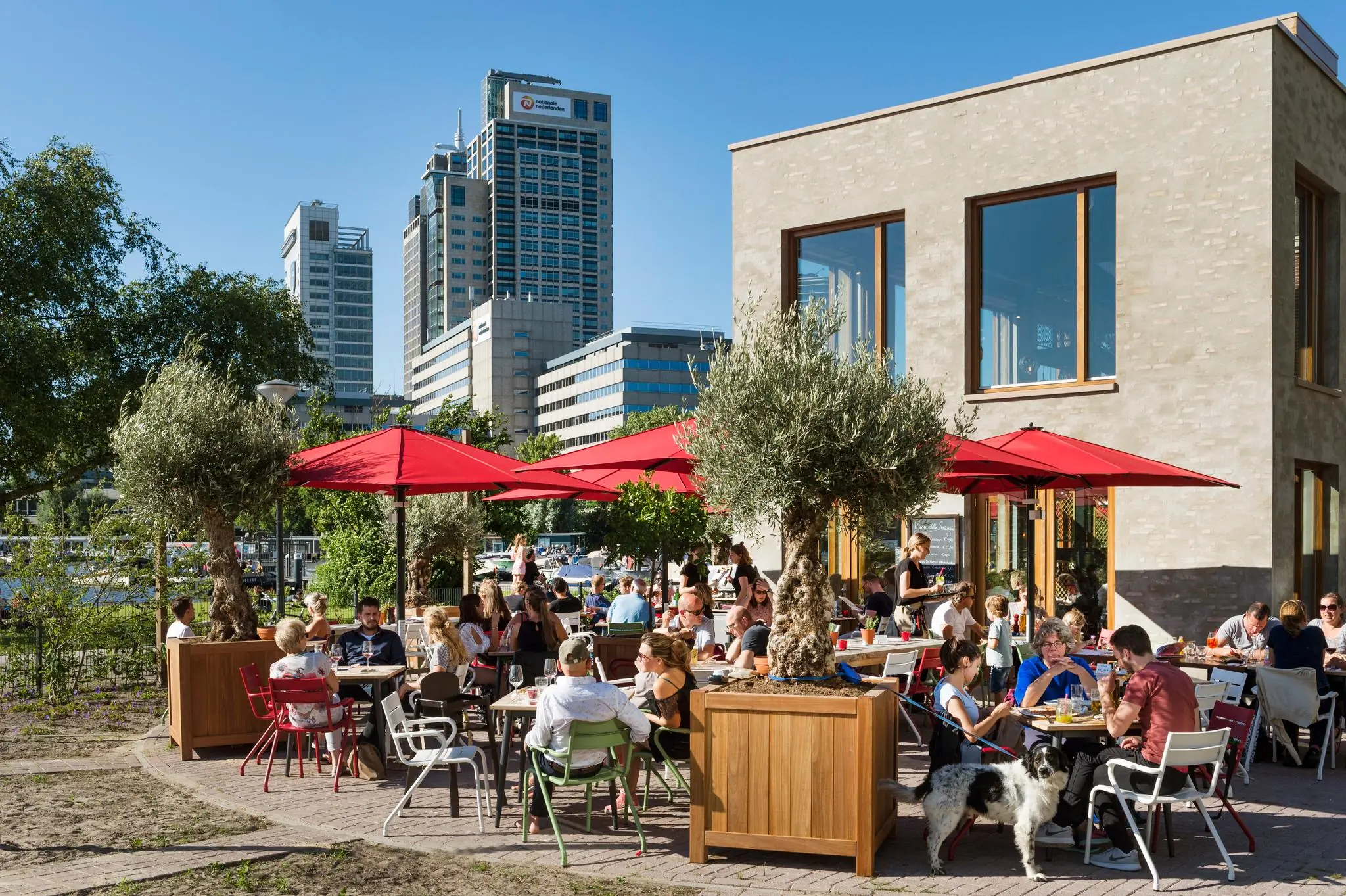 Terras met rode parasols en mensen die genieten van eten en drinken op het Amstelplein in Amsterdam, met kantoorgebouwen op de achtergrond.