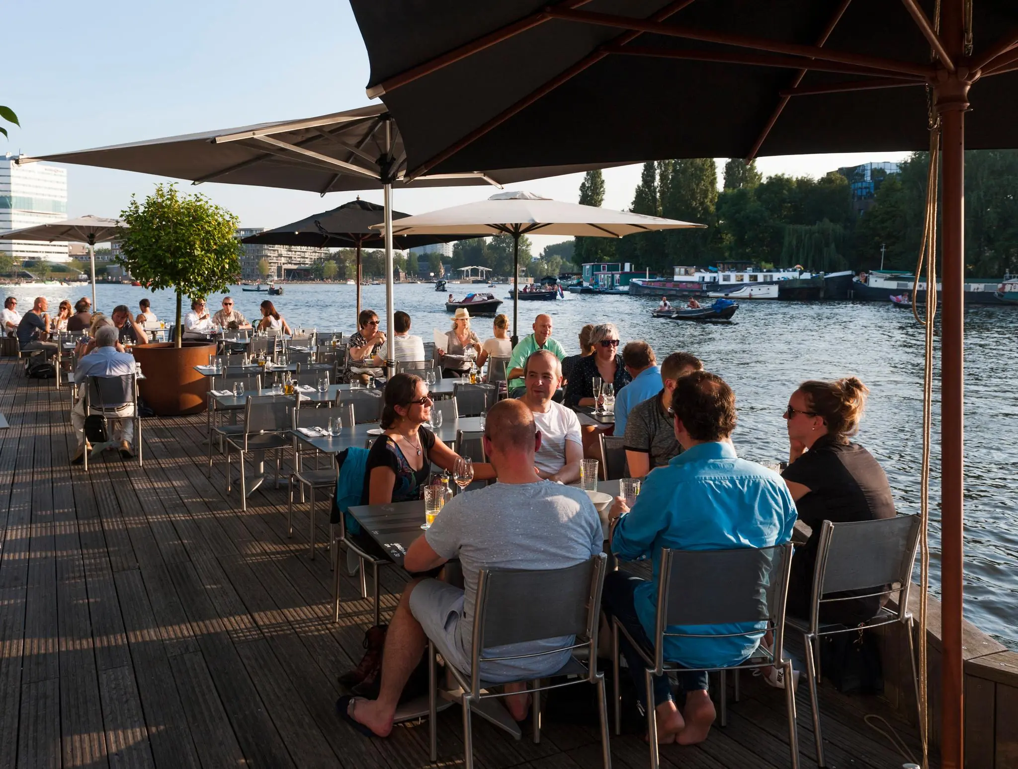 Terras aan het Amstelplein met mensen die genieten van eten en drinken aan het water op een zonnige dag.