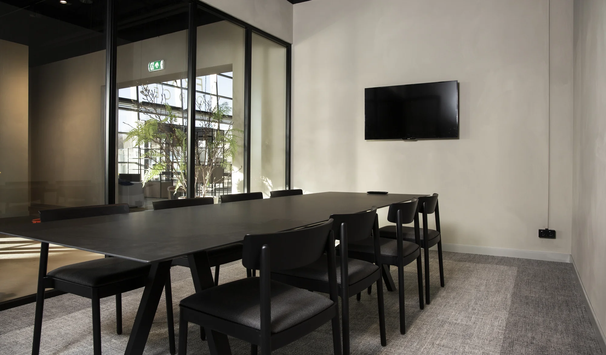 Moderne vergaderruimte aan de Atoomweg met zwarte tafel, acht stoelen, een wandtelevisie en glazen wand met uitzicht op planten.