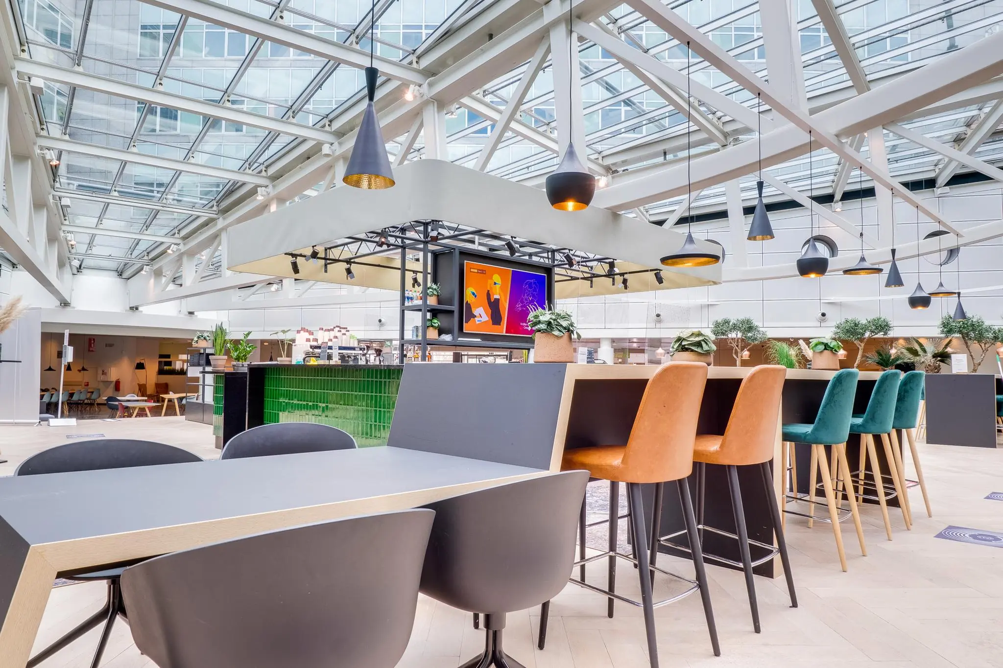 Moderne, lichtovergoten kantoorlounge aan de Winthontlaan met hoge tafels, kleurrijke barkrukken en hanglampen onder een glazen dak.