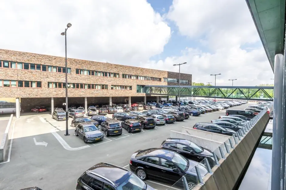 Parkeerterrein bij het Herculesplein met een groot kantoorgebouw en een glazen loopbrug op de achtergrond.