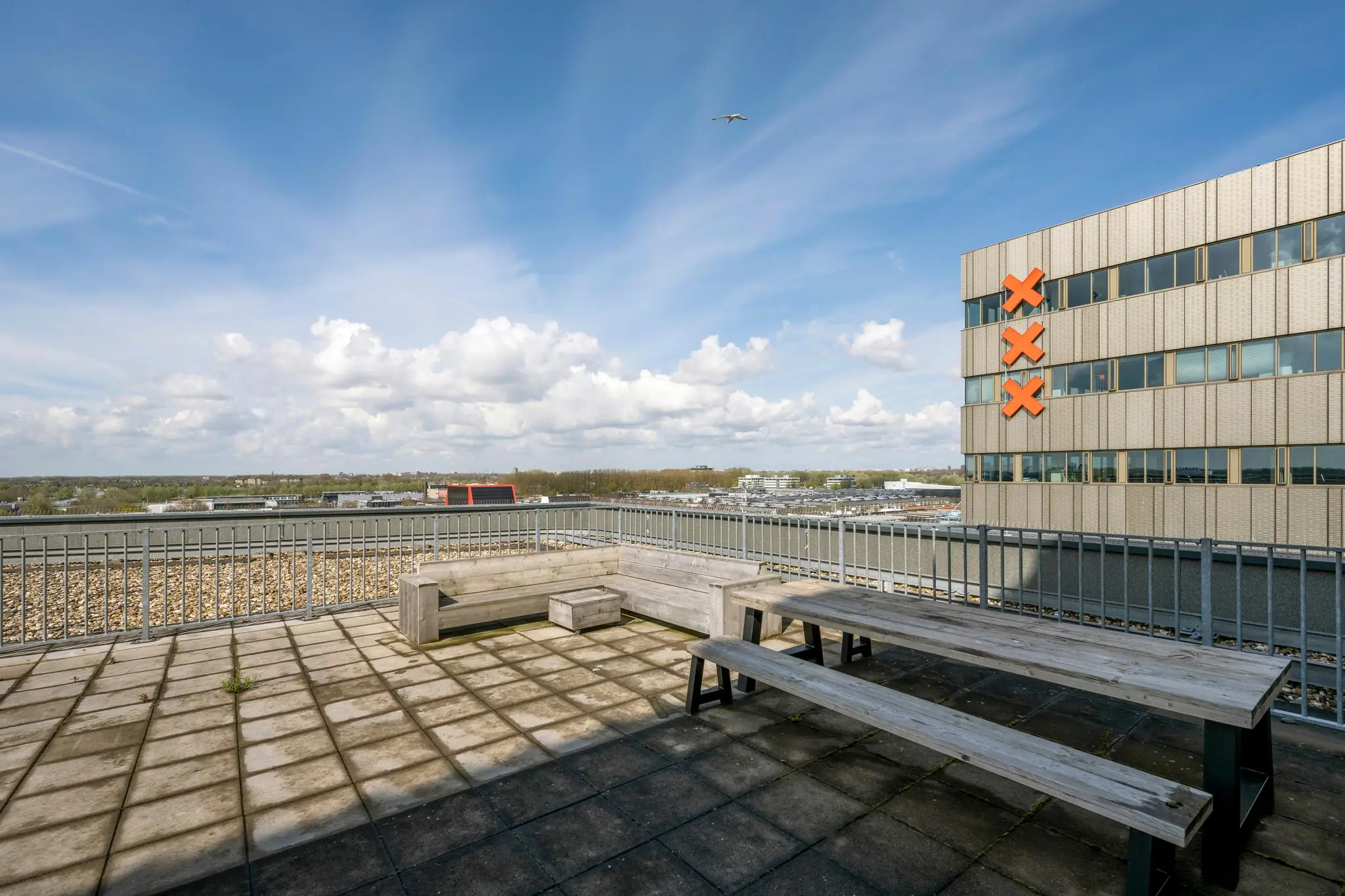 Zonnig dakterras met houten zitmeubilair en uitzicht op een kantoorgebouw aan de Herikerbergweg met drie rode Amsterdamse Andreas-kruisen.