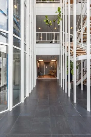 Licht en modern atrium met grote ramen, een betegelde vloer en een houten trap in een kantoorgebouw.