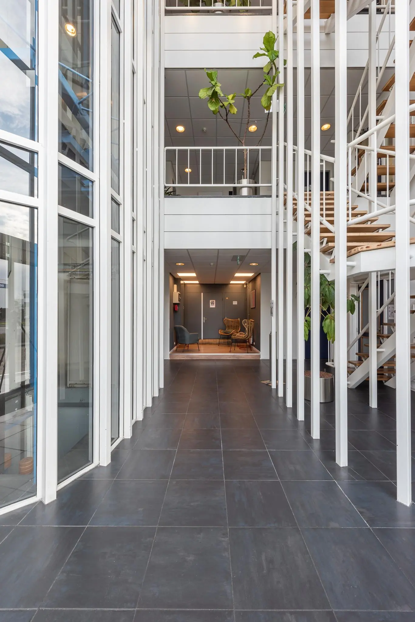 Licht en modern atrium met grote ramen, een betegelde vloer en een houten trap in een kantoorgebouw.