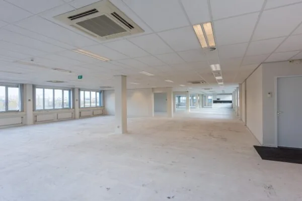 Ruime, lege kantoorruimte met systeemplafond, grote ramen en zichtbaar ventilatiesysteem.