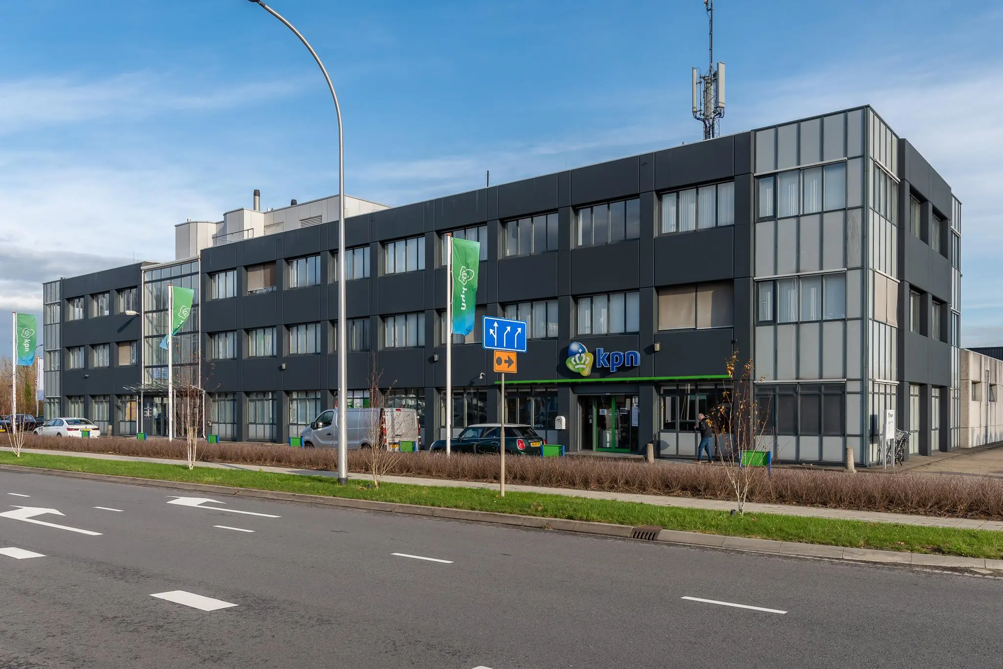 Voorgevel van een modern kantoorgebouw van KPN aan een brede weg met groenstrook en vlaggen.