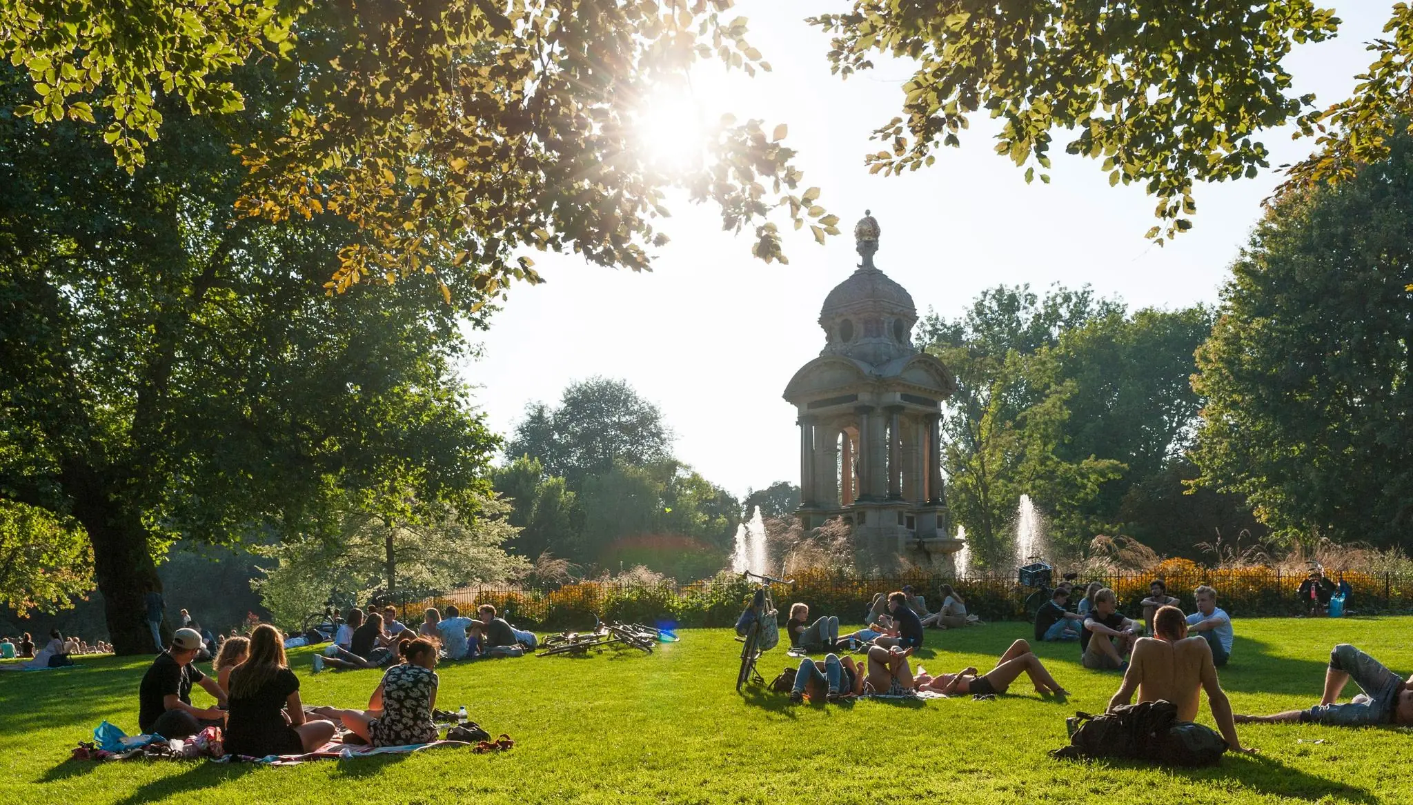 Mensen relaxen en picknicken in het zonnetje op een grasveld bij de fontein en het monument in het Vondelpark.