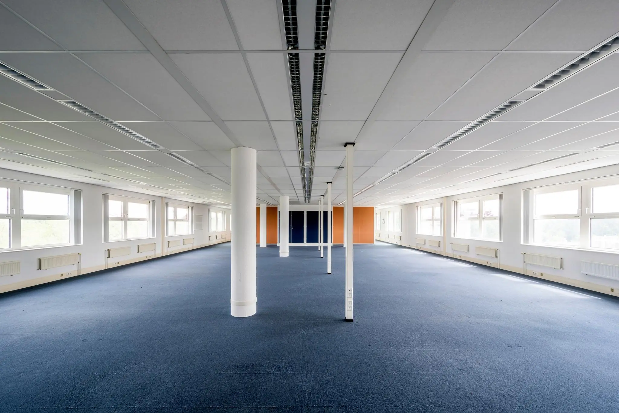 Lege kantooretage met blauw tapijt, witte pilaren en grote ramen aan beide zijden.