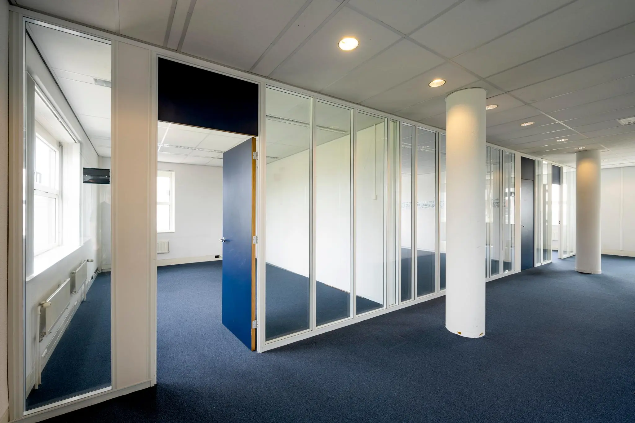 Moderne kantoorruimte met blauwe tapijtvloer, glazen wanden en een openstaande blauwe deur.
