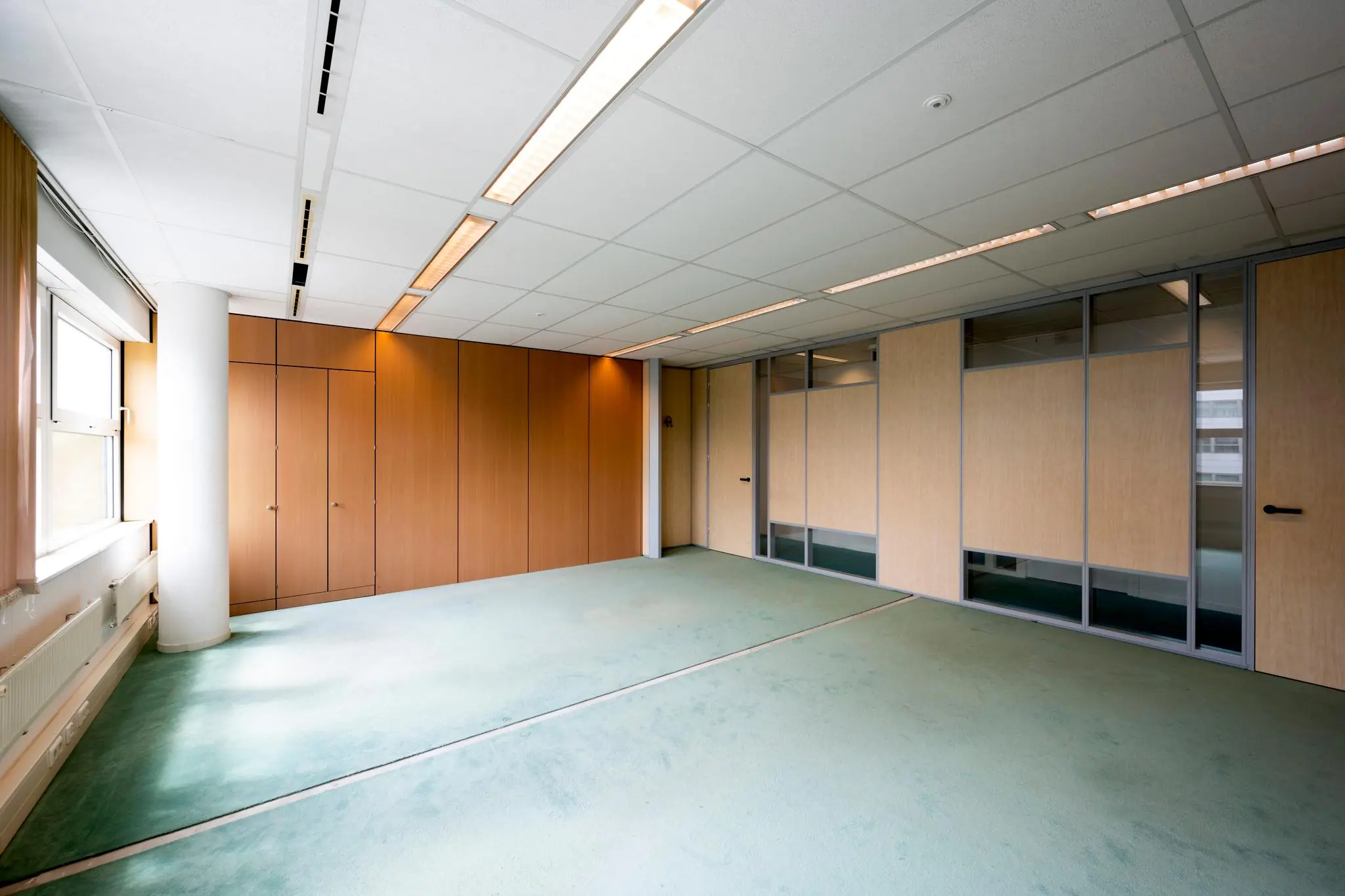 Lege kantoorruimte met groene vloerbedekking, houten wandpanelen en grote ramen.