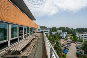 Buitenruimte met picknicktafels op een balkon van een modern kantoorpand, uitkijkend over een parkeerterrein en omliggende kantoorgebouwen.