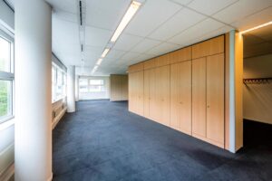 Lichte kantoorruimte met blauwe vloerbedekking, grote ramen en een wand met houten kasten.