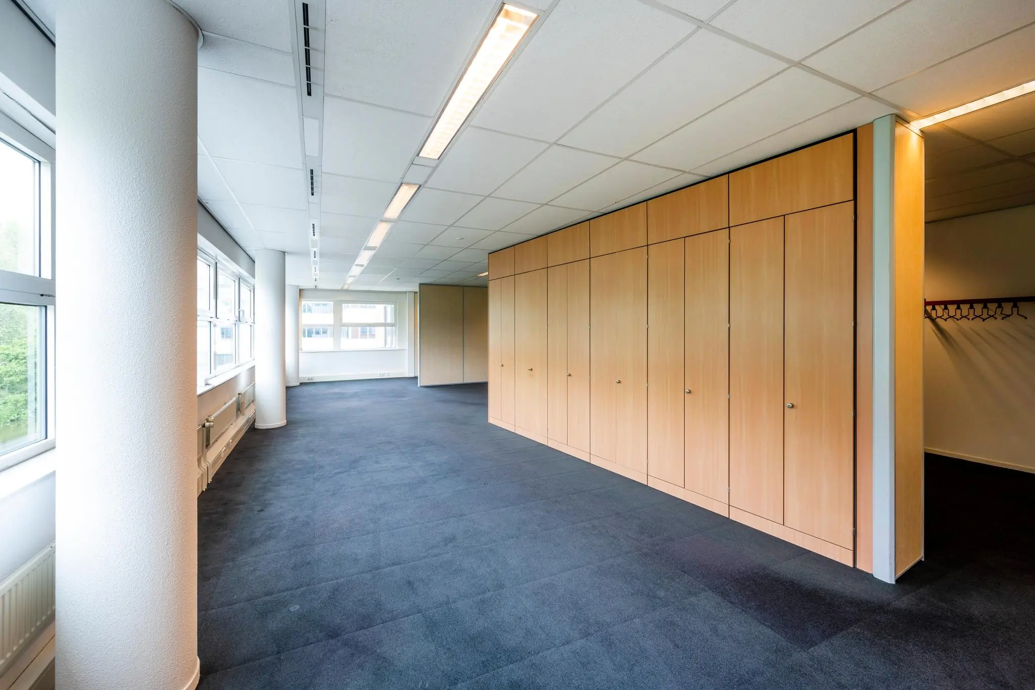 Lichte kantoorruimte met blauwe vloerbedekking, grote ramen en een wand met houten kasten.