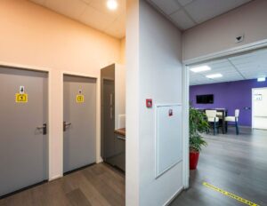 In een modern bedrijfsinterieur bevinden zich twee toiletten naast een pantry en een open ruimte met paarse muur en zitplaatsen.