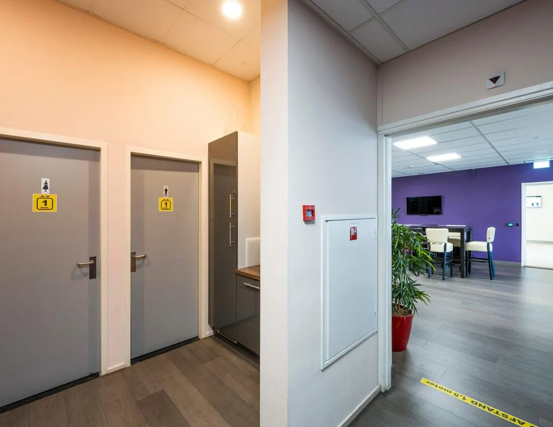 In een modern bedrijfsinterieur bevinden zich twee toiletten naast een pantry en een open ruimte met paarse muur en zitplaatsen.