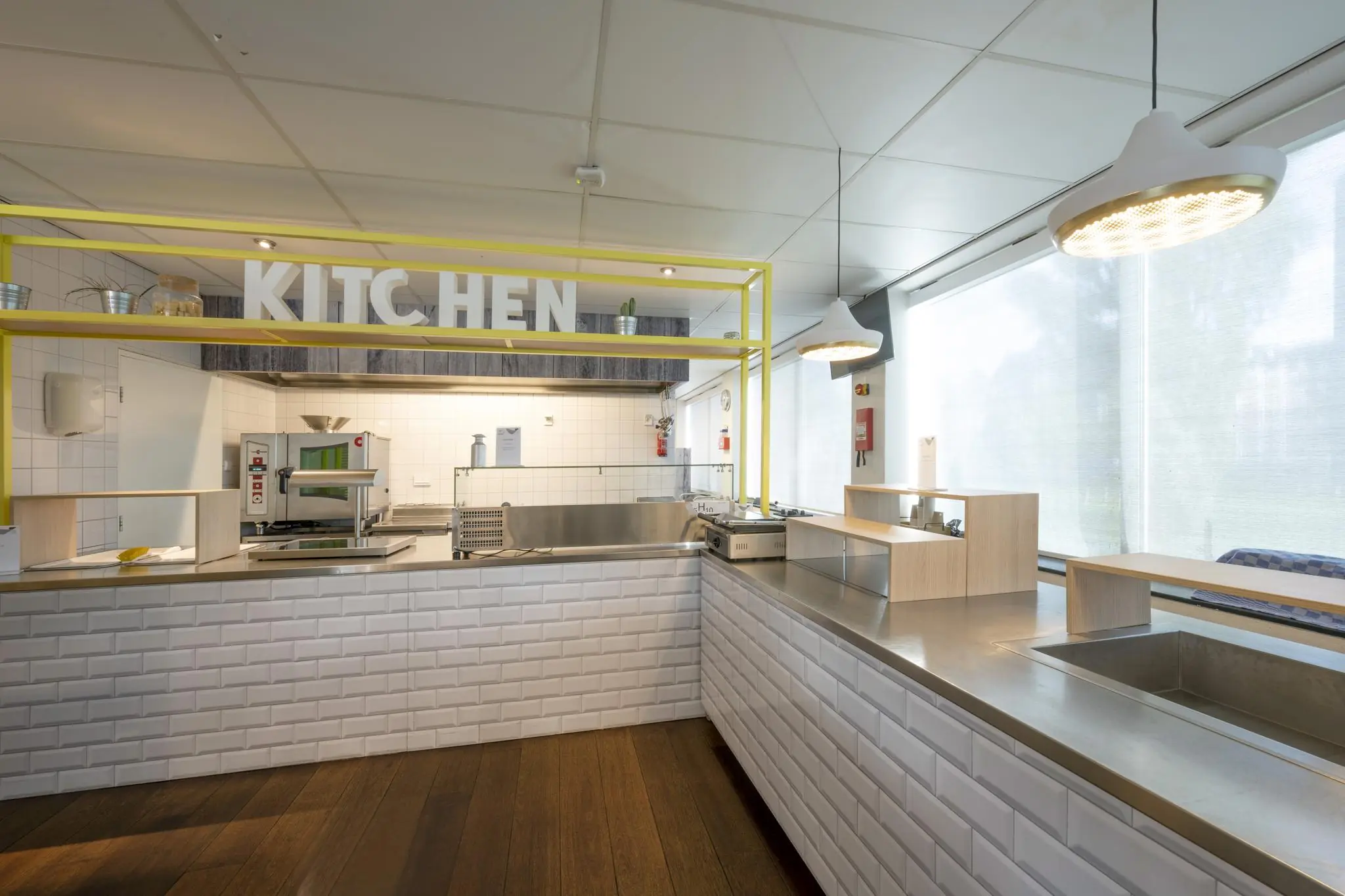 Moderne, lichte keuken met witte tegels, roestvrijstalen werkbladen en een gele omlijsting met het woord "KITCHEN" erop in Rietlandpark.