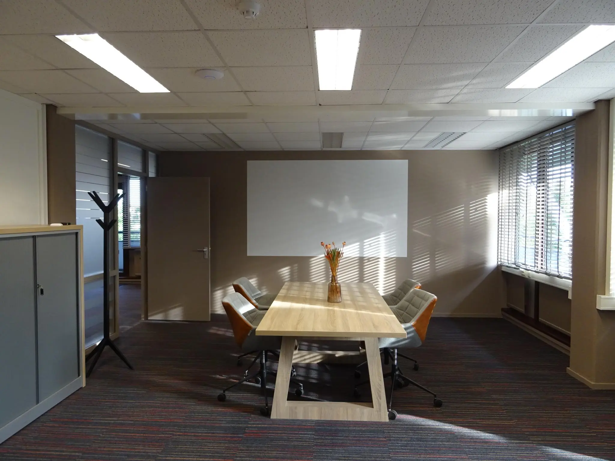 Vergaderruimte aan de Zaagmolenlaan met houten tafel, zes stoelen, whiteboard en zonlicht door jaloezieën.