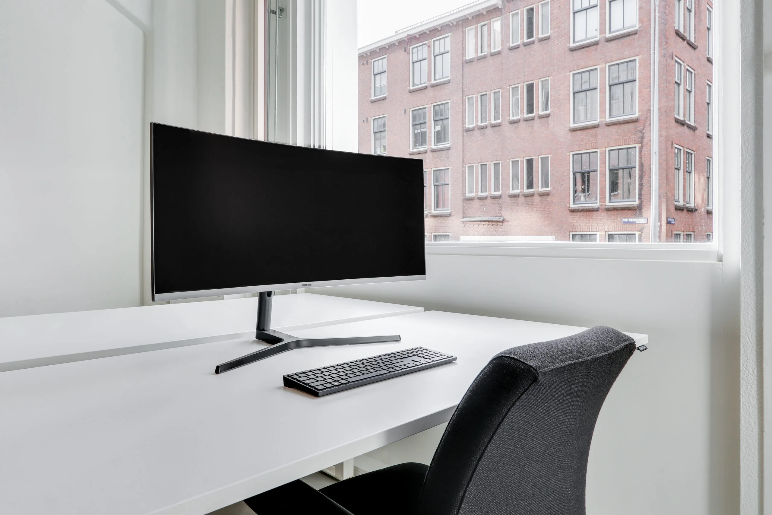 Minimalistisch co-working bureau in Amsterdam Houthavens met monitor, toetsenbord en uitzicht op bakstenen gevel.