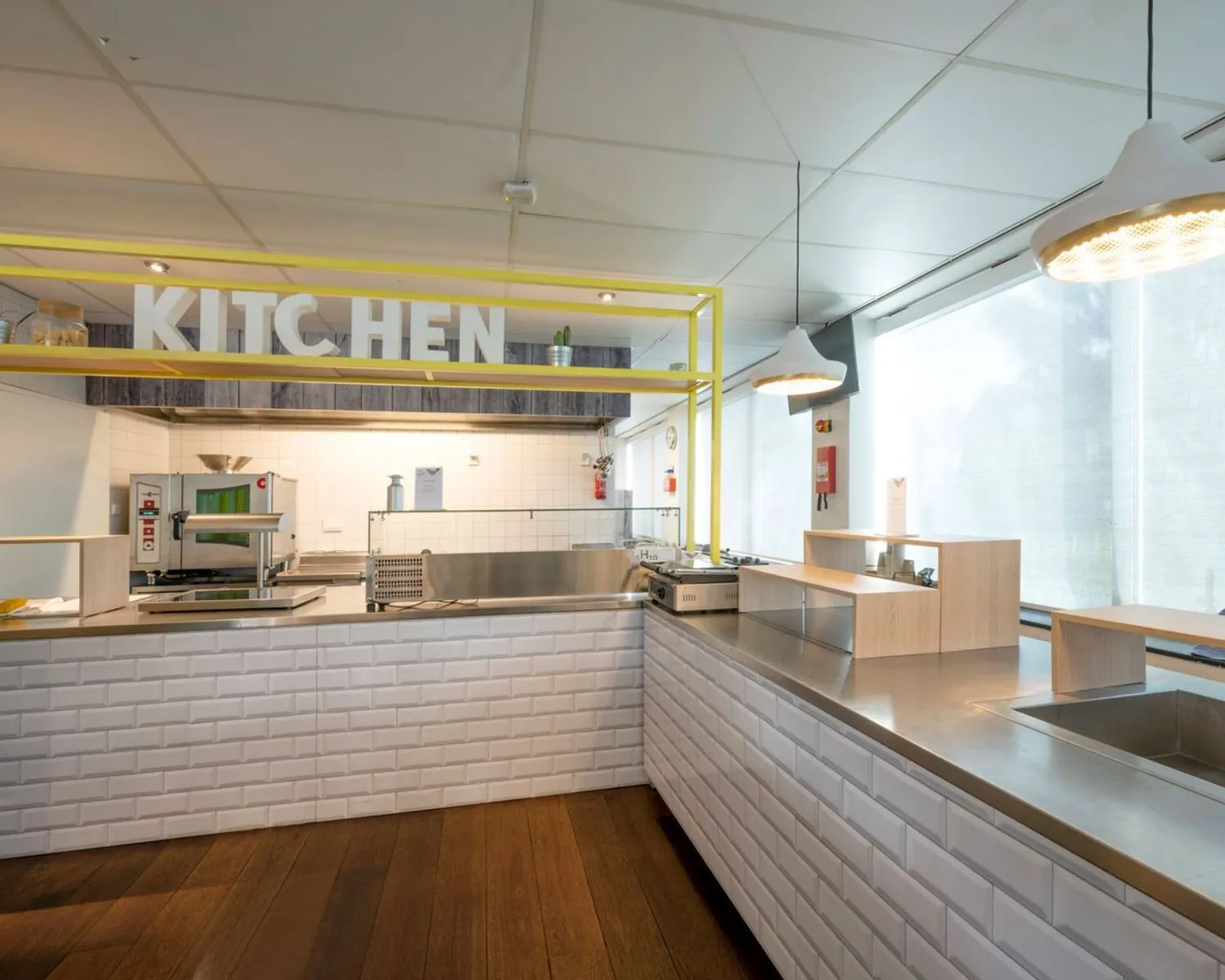 Moderne gedeelde kantoorkeuken met witte tegels, rvs werkbladen en opvallende gele accenten in Rietlandpark 309-357 te Amsterdam.