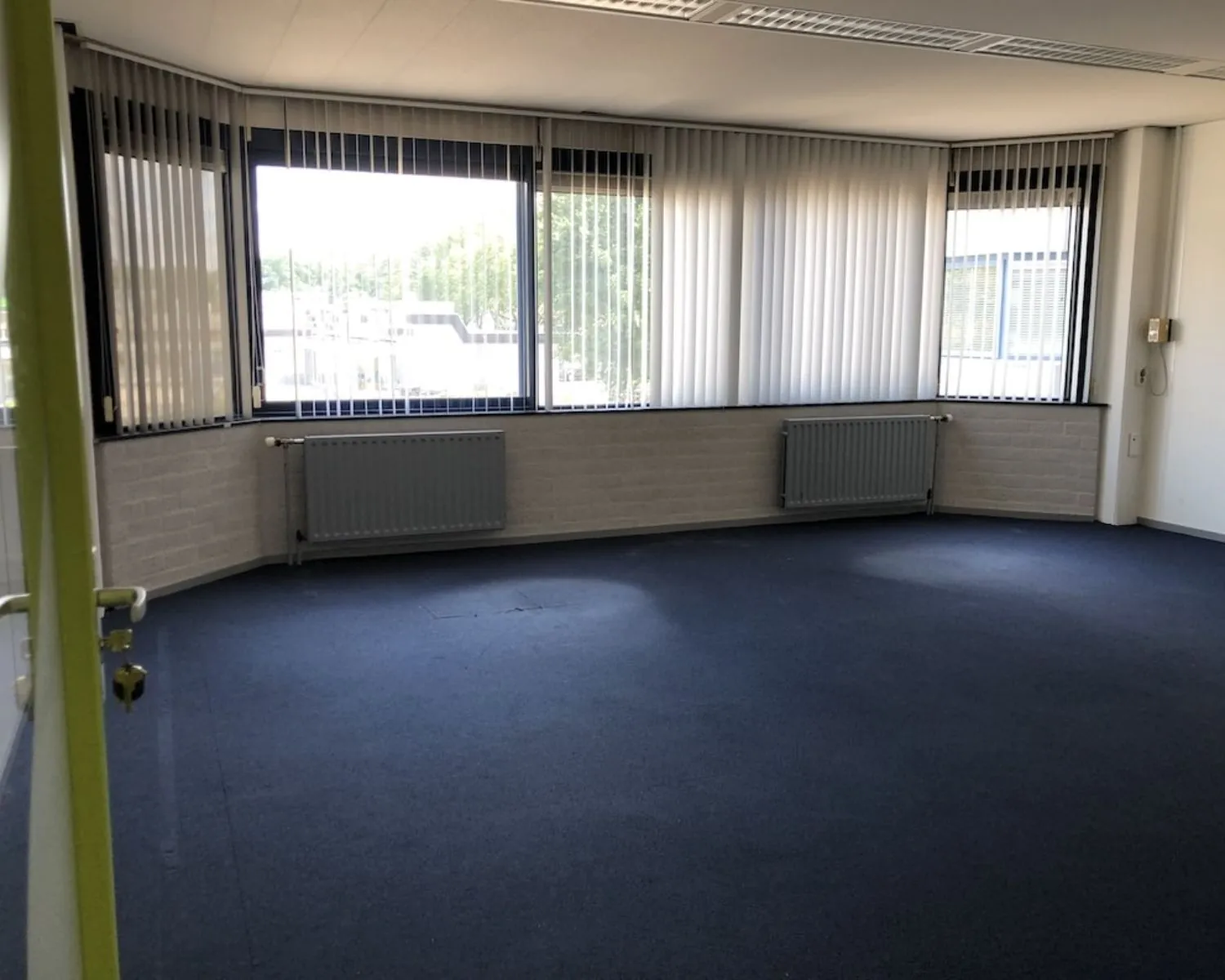 Lichte, lege kantoorruimte met blauw tapijt en grote ramen met verticale lamellen aan de Hooilaan 1 in Breda.